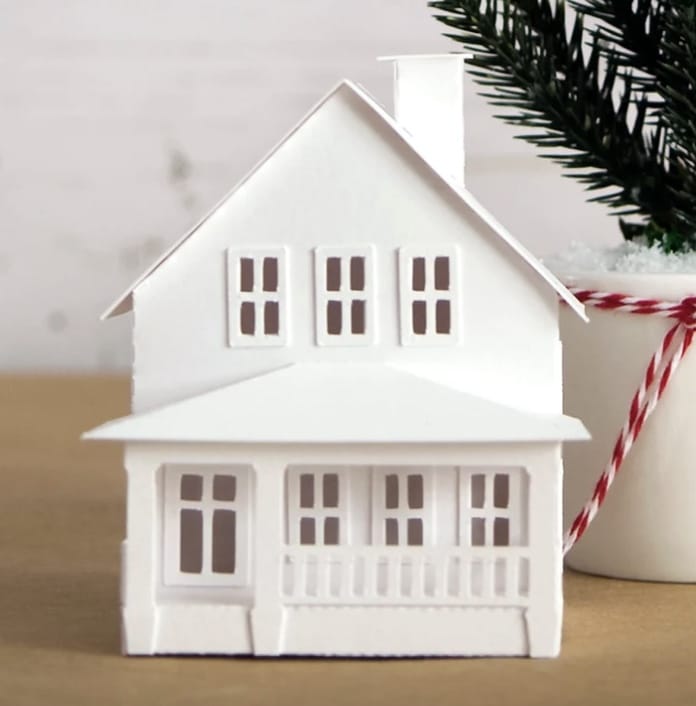 A small white paper model of a house with a porch and multiple windows rests on a wooden surface, embodying the essence of Christmas crafts for adults. Nearby, a white cup holds a small pine tree adorned with red and white string, completing the festive scene.